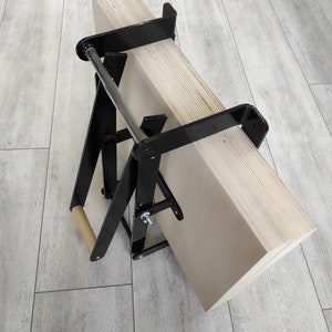 K&ouml;nnte beinhalten: Ein schwarzer Metallst&auml;nder mit einer Holzplanke, die mit zwei Klemmen befestigt ist. Der St&auml;nder hat einen Holzgriff und ist zum Halten und Transportieren gro&szlig;er Holzst&uuml;cke konzipiert.