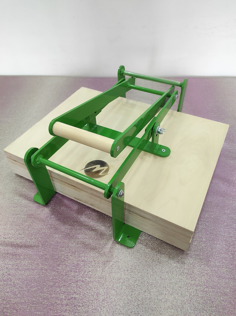 May include: Green metal press with a wooden handle, used to press together multiple pieces of wood. The press is sitting on a stack of plywood.