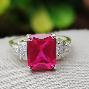 Peut inclure: Une bague en argent avec une grande pierre précieuse rose vif de forme carrée entourée de petites pierres précieuses claires.