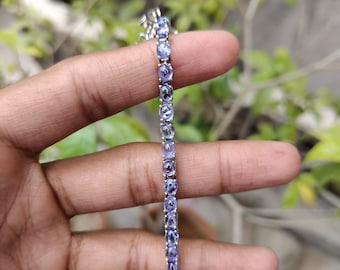 May include: A delicate bracelet featuring a series of oval-cut, light purple gemstones set in a silver-toned metal. The bracelet is held against a blurred background of green foliage, showcasing the jewelry's color and design.