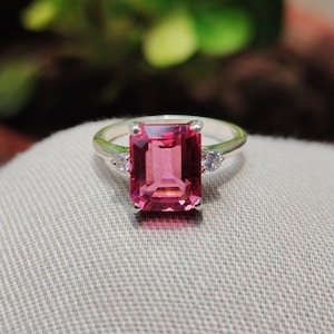 May include: A silver ring featuring a large, pink, emerald-cut gemstone and two smaller, white gemstones on the band.