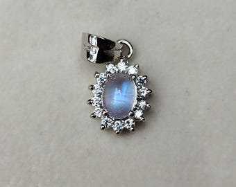 Può includere: Ciondolo in argento con una gemma ovale e iridescente, circondata da piccole gemme trasparenti. Il ciondolo ha un anello per una catena. Le gemme hanno una tonalità azzurra. È un gioiello.