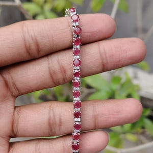 May include: A silver bracelet with a line of oval, deep red gemstones, interspersed with smaller, clear stones. The bracelet is displayed against a blurred green backdrop, highlighting the jewellery's sparkle.