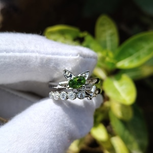 May include: A close-up of two silver rings. The top ring features a green gemstone and a butterfly design. The bottom ring has a series of small, clear gemstones. The rings are set against a blurred background of green leaves.