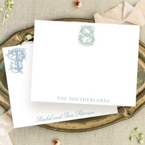 May include: Two white rectangular stationery cards featuring ornate, decorative monograms in muted blue and sage green. One card displays the text THE SOUTHERLANDS in gray, while the other features the script text Rachel and Ben Peterson in blue.