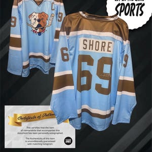 Puede incluir: Dos camisetas de hockey azul claro con detalles marrones y blancos. Una camiseta presenta un gráfico de bulldog con un palo de hockey. La otra camiseta tiene "SHORE" y "69" en la espalda. La imagen incluye un certificado de autenticidad y el texto "GET IN THE GAME SPORTS".