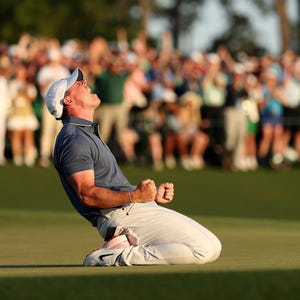Peut inclure: Un golfeur célèbre une victoire en s'agenouillant sur le green et en levant les bras en l'air. Il porte un chapeau blanc, une chemise bleue et un pantalon blanc. L'arrière-plan est flou et montre une foule de personnes qui applaudissent.