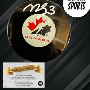 May include: A black hockey puck with white script reading "NS3". The puck has a circular emblem with a red maple leaf and the word "CANADA". A "Certificate of Authenticity" is included. The words "GET IN THE GAME SPORTS" are also visible.
