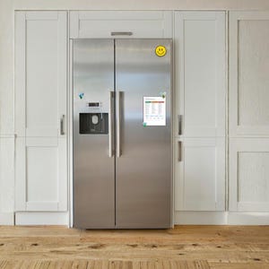 May include: A stainless steel side-by-side refrigerator with a water and ice dispenser on the left door. A yellow smiley face magnet is at the top, and a rectangular white chart with text and coloured rows is attached to the right door with magnets.
