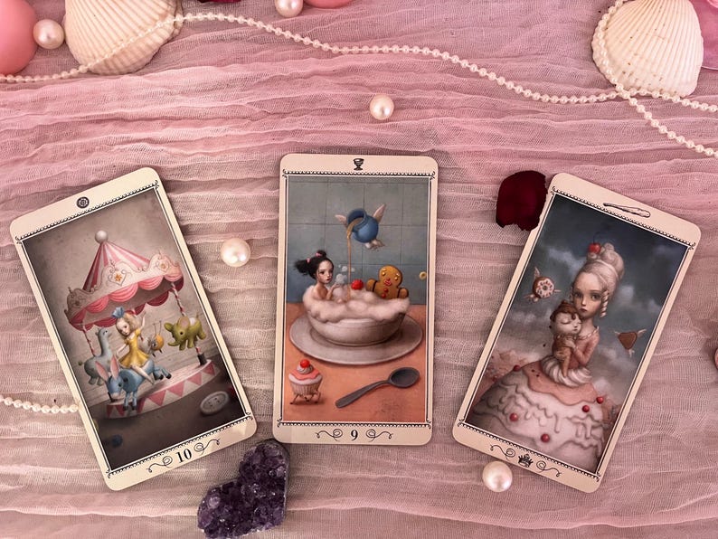 May include: Three rectangular tarot cards featuring whimsical illustrations. The left card shows a carousel with a figure, the center card depicts a figure in a bubble bath with a gingerbread man, and the right card shows a figure in a cake dress.