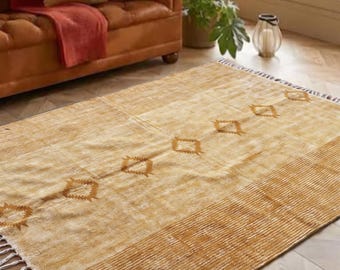 May include: A rectangular area rug in shades of mustard yellow and cream, featuring a central row of diamond-shaped geometric motifs, striped borders on the ends, and white fringe detailing along the short edges of the woven textile.