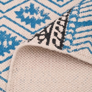 Op de afbeelding: Close-up van een wit tapijt met een blauw en zwart geometrisch patroon. Het tapijt is omgeslagen, waardoor de witte onderkant zichtbaar is.