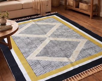 May include: A rectangular area rug featuring a black and white striped pattern with two large central diamond shapes containing a fine grid design. The rug is bordered by a mustard yellow band and a black edge, finished with beige fringe on the ends.