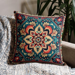 May include: Square throw pillow with a teal, red, and beige floral pattern. The pillow is placed on a gray sofa with a blue and white patterned throw blanket. A green plant is in the background.