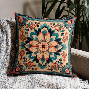 May include: Square decorative pillow with a floral design in teal, coral, and cream colors. The pillow is placed on a gray sofa with a blue and white patterned throw blanket. A green plant is in the background.