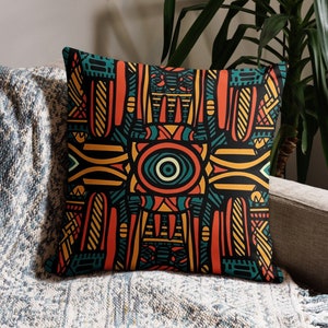 May include: Square throw pillow with a vibrant geometric pattern in orange, teal, yellow, and black. The design features abstract shapes and a central eye-like motif. The pillow is placed on a gray sofa with a blue and white blanket.