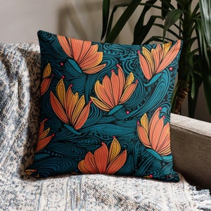 May include: Square throw pillow with a teal background and a repeating pattern of orange and yellow stylized flowers. The pillow is placed on a gray sofa with a blue and white patterned blanket.