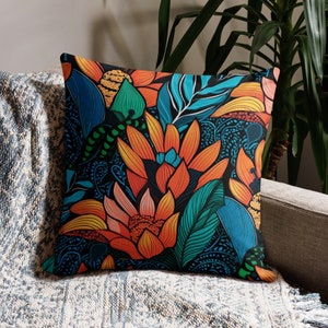 May include: Square throw pillow with a vibrant floral design. The pillow features large orange and yellow flowers, teal and green leaves, and a dark blue background. The pillow is placed on a gray sofa with a patterned throw blanket.