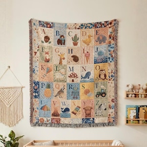 May include: A woven throw blanket featuring a grid of alphabet letters from A to Z, each paired with a corresponding illustration such as an acorn, bear, cactus, duck, elephant, fox, giraffe, hedgehog, ice cream, jellyfish, kite, lamb, mushroom, narwhal, owl, pineapple, quail, robot, sun, tractor, umbrella, volcano, whale, xylophone, yak, and zebra.