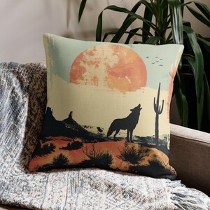 May include: A throw pillow with a desert landscape design. The pillow features a silhouette of a wolf howling at a large orange sun, with a cactus in the background. The pillow is beige with orange, brown, and green accents.