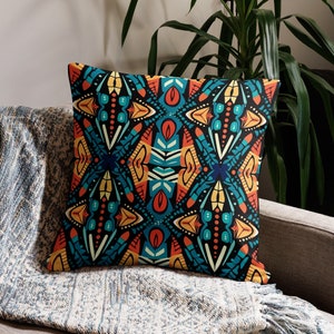 May include: Square throw pillow with a vibrant, symmetrical pattern in teal, orange, yellow, and red on a black background. The pillow is resting on a gray sofa with a blue and white patterned throw blanket.