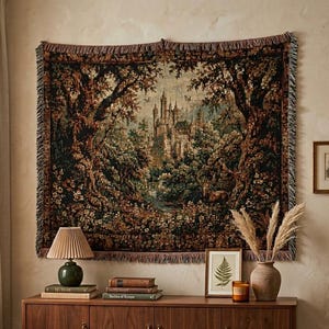 Puede incluir: Un tapiz de pared tejido rectangular que presenta una escena de paisaje detallada con un castillo central rodeado de &aacute;rboles densos, follaje y flores en tonos verde oscuro, marr&oacute;n y dorado, acabado con un borde con flecos en los cuatro lados.