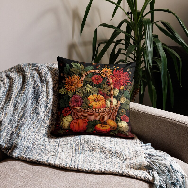 Puede incluir: Un coj&iacute;n cuadrado con fondo oscuro que muestra una cesta de mimbre llena de calabazas naranjas, girasoles amarillos y dalias de color rojo intenso. El dise&ntilde;o incluye follaje verde y peque&ntilde;as calabazas dispuestas alrededor de la cesta.