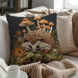May include: Decorative throw pillow featuring an embroidered hedgehog surrounded by mushrooms and flowers. The pillow has a dark gray background and is placed on a cream-colored sofa. The hedgehog has brown and tan quills, with orange and white mushrooms.