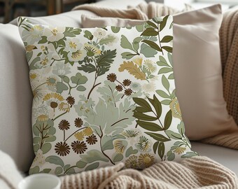 May include: A throw pillow with a white background and a floral pattern in shades of green, brown, and yellow. The pillow is sitting on a couch with a brown knitted throw blanket.