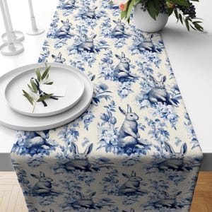 May include: A blue and white table runner with a repeating pattern of bunnies and flowers. The runner is made of a soft, woven fabric and is perfect for adding a touch of spring to your table.