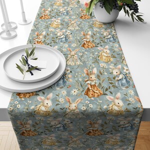 May include: A light blue table runner featuring a repeating pattern of bunnies in dresses and floral accents. The bunnies are in various dresses of yellow, blue, and brown. White plates and candles are on the table.