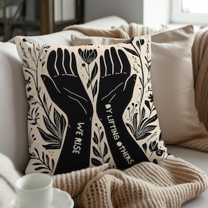 May include: A beige throw pillow with a black and white design of two hands reaching up with the text "We Rise By Lifting Others" in black lettering.