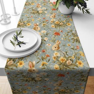 May include: A light blue table runner with a floral and bunny rabbit design. The runner features yellow, orange, and white flowers, with several bunny rabbits wearing dresses. The runner is placed on a white table, with white plates and candles.