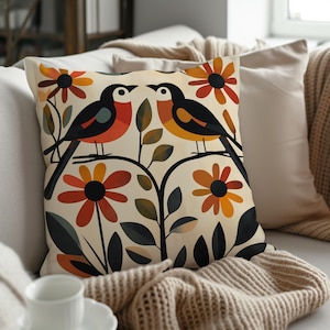 May include: A throw pillow with a cream background featuring two stylized birds perched on a branch with orange, red, and yellow flowers. The birds are facing each other and have black, orange, yellow, and red accents.