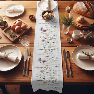 May include: A white table runner with a floral pattern featuring a variety of colorful wildflowers. The runner is laid out on a wooden table with place settings on either side.