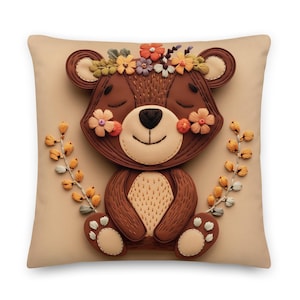 May include: A decorative square pillow featuring a smiling brown bear with a floral crown and blush-colored cheeks. The bear is surrounded by embroidered floral accents and sits on a beige background. The pillow is a charming addition to any room.