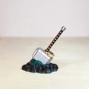 May include: A silver and brown Mjolnir hammer ring holder with a gold ring on top, sitting on a black and teal rock base.