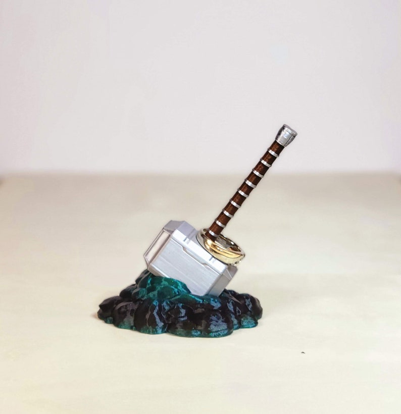 May include: A silver and brown Mjolnir hammer ring holder with a gold ring on top, sitting on a black and teal rock base.
