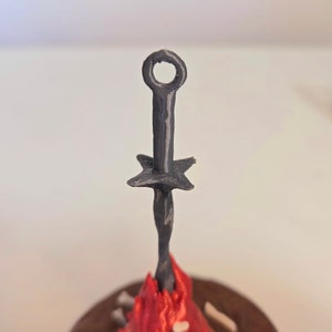 Puede incluir: Una hoguera en miniatura negra, impresa en 3D, con una espada. La espada tiene un anillo en la parte superior y una guarda en forma de estrella. Las llamas son rojas y naranjas, y la base es marr&oacute;n con detalles de huesos.