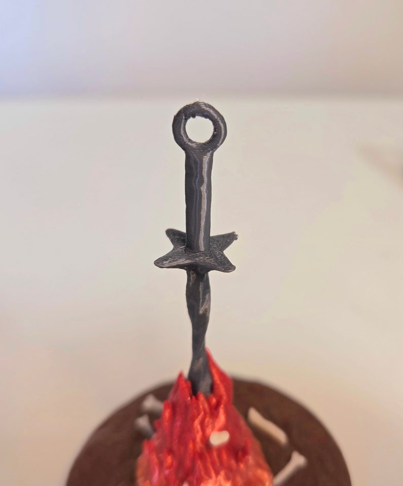 Puede incluir: Una hoguera en miniatura negra, impresa en 3D, con una espada. La espada tiene un anillo en la parte superior y una guarda en forma de estrella. Las llamas son rojas y naranjas, y la base es marr&oacute;n con detalles de huesos.