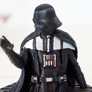 May include: A black plastic figurine of Darth Vader from Star Wars. The figurine is wearing a black cape and has a white stripe across the chest. The figurine is holding its right hand up in a gesture of greeting.