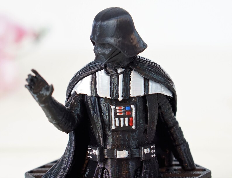 May include: A black plastic figurine of Darth Vader from Star Wars. The figurine is wearing a black cape and has a white stripe across the chest. The figurine is holding its right hand up in a gesture of greeting.