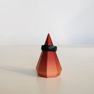 May include: A black silicone ring displayed on a red and orange geometric ring holder.