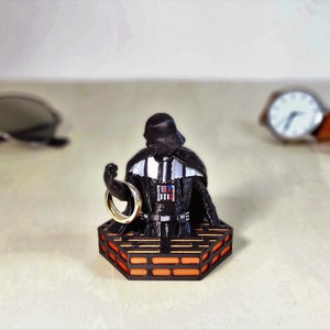 May include: A black Darth Vader figurine holding a gold ring, standing on a hexagonal platform with orange and black details.