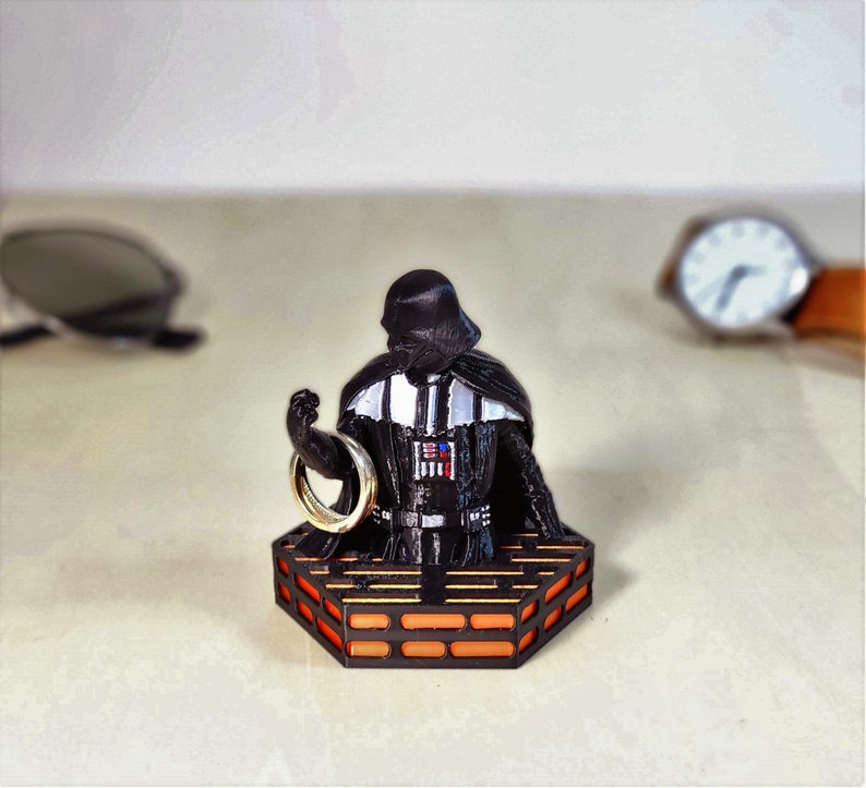 May include: A black Darth Vader figurine holding a gold ring, standing on a hexagonal platform with orange and black details.