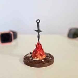 Puede incluir: Una escultura en miniatura de una hoguera con una espada. La base es marr&oacute;n, con detalles blancos en forma de hueso. Las llamas son rojas y naranjas, y la espada es negra. La escultura est&aacute; sobre una superficie blanca.