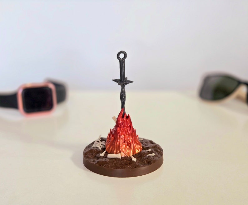 Puede incluir: Una escultura en miniatura de una hoguera con una espada. La base es marr&oacute;n, con detalles blancos en forma de hueso. Las llamas son rojas y naranjas, y la espada es negra. La escultura est&aacute; sobre una superficie blanca.