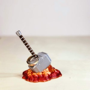 May include: A silver Mjolnir ring holder with a brown handle, sitting on a red and orange base. Two gold rings are placed on the top of the holder.