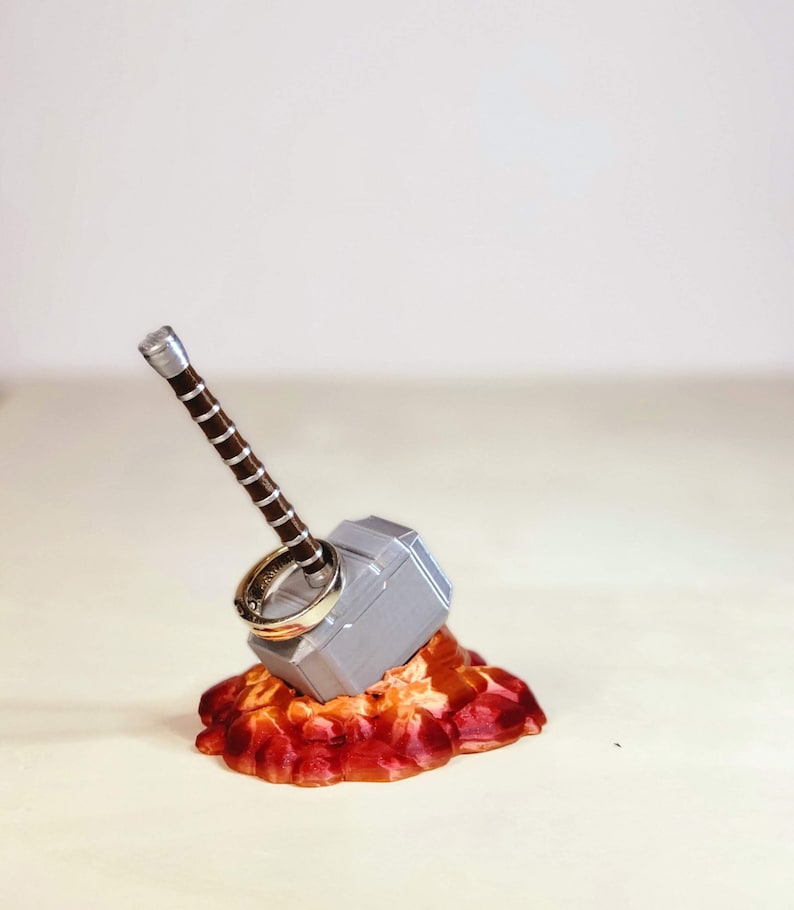 May include: A silver Mjolnir ring holder with a brown handle, sitting on a red and orange base. Two gold rings are placed on the top of the holder.