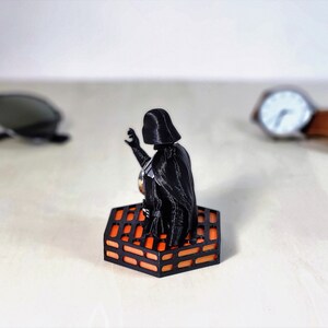 May include: A black Darth Vader figurine standing on an orange and black hexagonal base. The base is designed to look like the Death Star trench run.